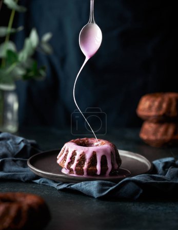 Téléchargez les photos : Mini gâteau bundt au chocolat avec glaçure rose étant versé sur elle à partir de la cuillère. Torse de personne dans les coulisses. Fond gris foncé et table en pierre noire. Style de vie image verticale avec un élément humain. - en image libre de droit