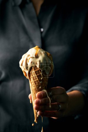 Photo for Woman's hand holding a cone of sea salt caramel ice cream. Ice cream is melting. She is wearing dark grey blouse. Vertical image with space for text. Light falling on the ice cream making in hero of the shot. - Royalty Free Image