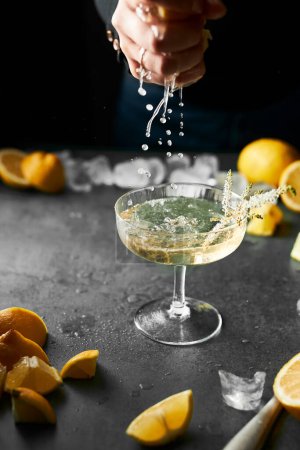Photo for Cropped hand squeezing lemon juice to a glass of see-through yellow cocktail. Lots of lemons. Moody still life. Refreshing drink. Grey color stone counter top. - Royalty Free Image