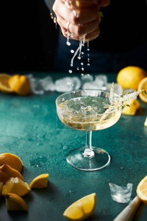Photo for Cropped hand squeezing lemon juice to a glass of see-through yellow cocktail. Lots of lemons. Moody still life. Refreshing drink. - Royalty Free Image