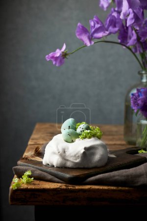 Photo for Small meringue pavlova topped with tiny easter eggs on a rustic wooden table. Festive easter still life photography. Dark grey background. Vertical lifestyle image. - Royalty Free Image