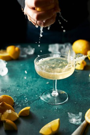 Photo for Cropped hand squeezing lemon juice to a glass of see-through foamy yellow cocktail. Lots of lemons. Moody still life. Refreshing drink. - Royalty Free Image