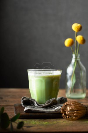 Photo for A glass of matcha latte on a rustic wooden table and grey background. Indoors set up. There are flowers, napkin, cutting board and bamboo whisk on the table. Vertical lifestyle image. - Royalty Free Image