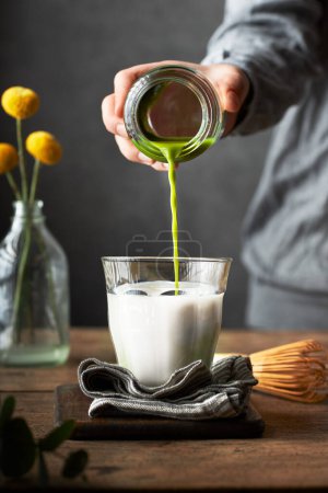 Téléchargez les photos : Découpé main versant coup de matcha dans un verre de lait pour préparer matcha latte. Table en bois et fond gris. Cadre salon ou salle à manger - en image libre de droit