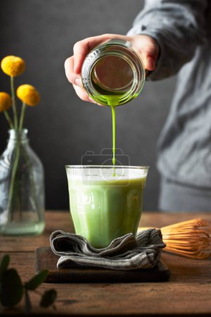 Photo for Cropped out hand pouring matcha to a glass of milk. Preparation of matcha latte. Wooden table and grey background. Living room or dining room setting. - Royalty Free Image