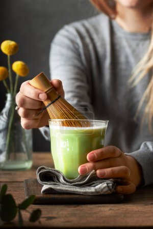Photo for Woman stirring matcha tea with bamboo whisk holding the glass. Grey background and wooden table. Living room or dining room home location. - Royalty Free Image
