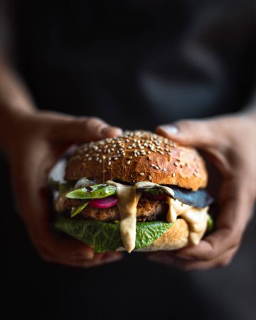 Photo for Vegetarian burger with vegan patty, asperges, pickled radishes and delicious dripping yellow sauce being held in the hands of a woman with her dark grey apron as a background. Vertical close up image. - Royalty Free Image