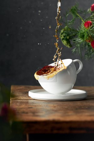 Photo for Artistic image of pouring black tea in a cup. Tea is splashing around. Cup is partly in the air. Dark background and rustic old wooden table. Flowers in the background and foreground. Vertical image. - Royalty Free Image