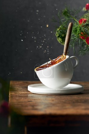 Photo for Artistic image of splashing black tea in a white classic cup which is standing partly in the air. Dark background and rustic old wooden table. Flowers in the background and foreground. Vertical image. - Royalty Free Image