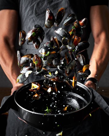 Photo for Preparation image of mussels. Male's hands throwing mussels in the air from the black pot to stir them around. There are drops around. Dark black background. - Royalty Free Image