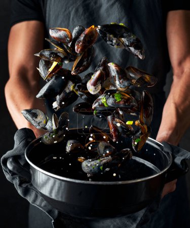 Photo for Preparation image of mussels. Male's hands throwing mussels in the air from the black pot to stir them around. There are drops around. Dark black background. - Royalty Free Image