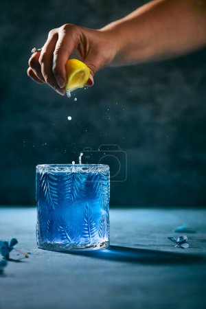Photo for Dark blue cocktail. Cropped hand squeezing lemon juice in the cocktail. Dark blue table and flowers on it. Black background. Vertical lifestyle image. - Royalty Free Image