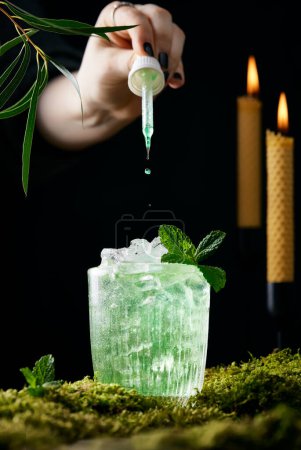 Téléchargez les photos : La main coupée goutte à goutte dans un cocktail vert clair très glacé. Cocktail debout sur la mousse, fond noir, bougies allumées. Images de style de vie vertical avec une atmosphère mystérieuse. - en image libre de droit