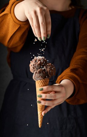Foto de Mujer rociando sal marina encima de helado de chocolate negro. Ella sostiene el cono de helado con su otra mano. Su torso es un fondo para la imagen. Estilo de vida oscuro imagen vertical. - Imagen libre de derechos