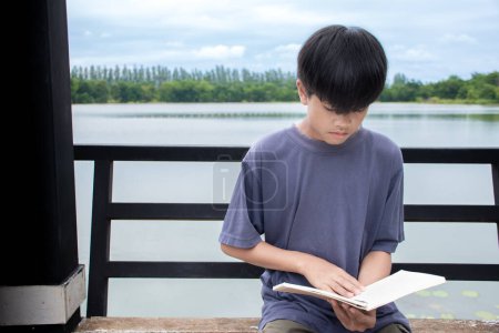 Téléchargez les photos : Asiatique jeune garçon lecture livre sur chaise au bord de la rivière fond est forêt et ciel avec lever de soleil, garçon chemise bleue, enfants avec livre, concept learning et éducation, naturel beau fond. - en image libre de droit
