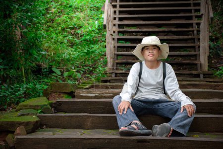 Asiatischer Junge mit weißem Hemd und langer Hose und Wanderhut sitzt auf den hölzernen Stufen, die bei natürlichem Wald- und Sonnenlicht den Berg hinauf führen.