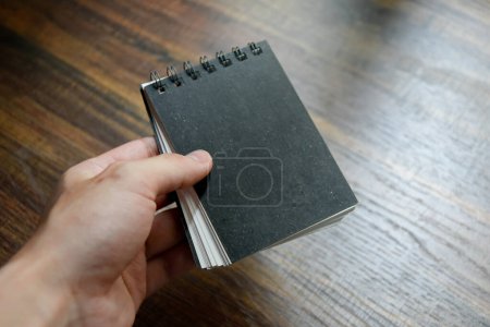 Téléchargez les photos : Main d'homme d'affaires tenant un petit carnet noir sur table de bureau motif en bois au bureau. - en image libre de droit