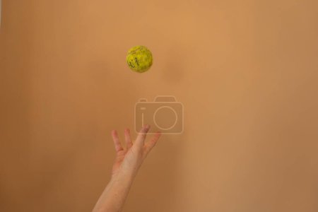 Téléchargez les photos : Balle de tennis vintage à lancer à la main contre le mur de jacquier : Une main lançant une balle de tennis antique dans l'air gelé dans le temps, sur fond de mur peint dans les tons de jacquier - en image libre de droit