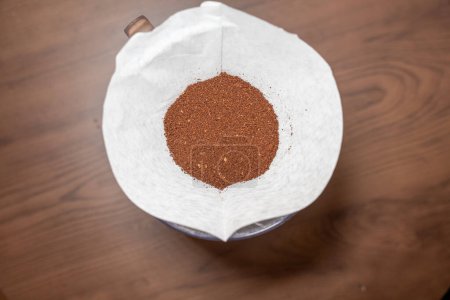 A white coffee filter nestled atop a V60 container, brimming with freshly ground coffee against the backdrop of a wooden table