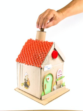 Hand putting coins into a unique house-shaped piggy bank made from recycled materials. Concept about saving and finance.