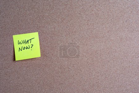 The words What Now on a yellow sticky note posted on a corkboard