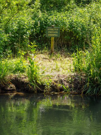 Una orilla del río serena con un follaje verde alto y un cartel que indica 'estrictamente pesca privada' cerca del borde del agua.