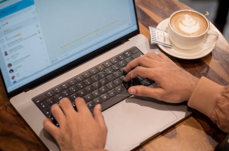 Mumbai, Indien 7. Dezember 2022 Ein Mann tippt in einem Chat auf Laptop und Kaffeetasse und Untertasse auf Holztisch beiseite gehalten