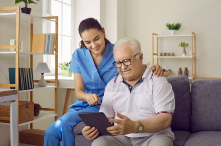 Photo for Young pretty brunette nurse at home communicates with an elderly man with glasses, showing him something on a tablet, conveying good mood and positive attitude. The concept of psychological health. - Royalty Free Image