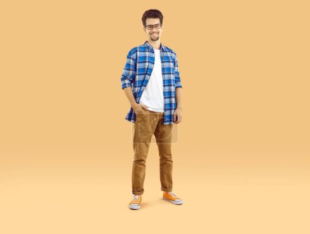 Téléchargez les photos : Plan studio d'un jeune homme heureux en tenue décontractée. Étudiant homme en T-shirt, chemise à carreaux bleue, pantalon marron et lunettes debout la main dans la poche sur un fond beige clair. Concept de mode - en image libre de droit