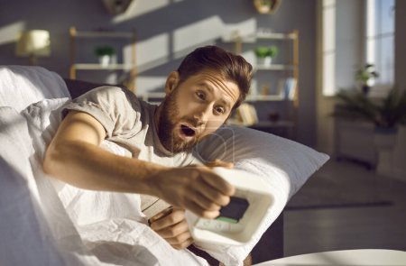 Photo for Emotional shocked overslept young redhead man holding alarm clock in his hand, waking up too late for work lying in bed at home. Oversleeping and bad morning concept. Selective focus - Royalty Free Image