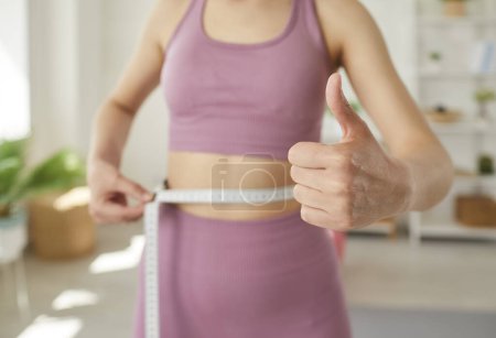 Photo for Cropped photo of faceless sporty slim woman wearing sportswear measure the waist with measuring tape in gym or at home and showing thumb up sign. Diet, slimming and healthy lifestyle concept. - Royalty Free Image