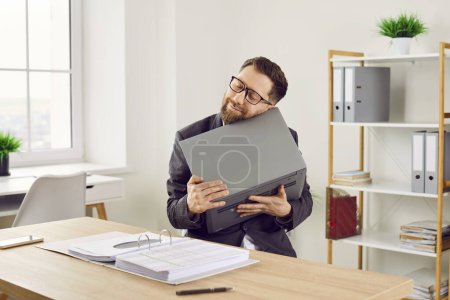 Photo for Happy, funny, smiling financial accountant loves his laptop very much. Young man sitting at office desk with business paperwork and tenderly holding notebook computer in his hands - Royalty Free Image