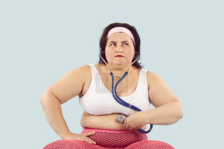 Photo for Obese large chubby overweight woman in sports clothes holds stethoscope on skin of big fat belly, listens to bowel sounds and thinks of digestive system health, overeating and stomach problems - Royalty Free Image