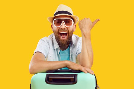 Photo for Portrait of young happy funny man tourist in sunglasses and beach hat with suitcase pointing index finger to the side on yellow background. Vacation advertising, summer holiday and travel concept. - Royalty Free Image