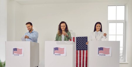 Téléchargez les photos : Les électeurs américains au bureau de vote lors des élections présidentielles. Groupe de trois personnes souriantes et heureuses debout dans des isoloirs blancs avec des drapeaux américains. Elections aux Etats-Unis concept - en image libre de droit