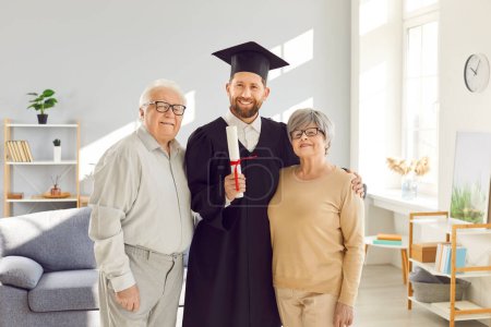 Photo for Portrait of happy young man student in university graduate gown and diploma in hands standing with senior old parents at home and looking proudly at camera. Graduation and education concept. - Royalty Free Image
