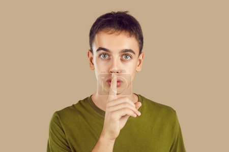 Photo for Teenage boy doing silencing Shh gesture. Studio headshot of adolescent boy in casual khaki green T shirt looking at camera with serious face, holding finger on lips, and asking you to keep his secret - Royalty Free Image