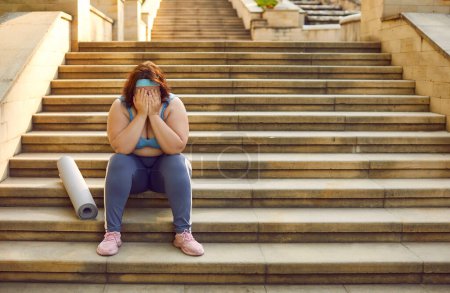Sad tired fat chubby overweight woman in sportswear with fitness mat sitting on stone steps outside gym, holding face in hands, feeling defeated, crying like loser. Sport, weightloss, failure concept