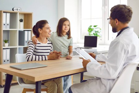 Mother and child visit family doctor at modern clinic. Happy, smiling mom and daughter sitting at table together with man pediatrician and talking about medical checkup results. Healthcare concept 