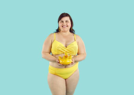 Photo for Portrait of a funny overweight smiling fat woman looking at camera wearing yellow bikini holding inflatable duck isolated on studio blue background. Summer holiday trip and vacation concept. - Royalty Free Image
