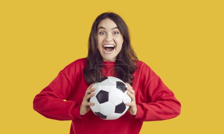 Téléchargez les photos : Portrait d'une belle femme brune super excitée vêtue d'un pull rouge chaud tenant un ballon de football. Photo studio d'un ardent fan de football sur fond jaune isolé. Concept de jeux de sport. - en image libre de droit