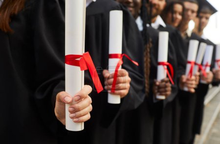 Photo for Hands of a graduate students standing in a row in black robes with diplomas in their hands outdoor at graduation ceremony close-up, selective focus. Graduating from university or college concept. - Royalty Free Image