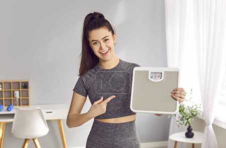 Retrato de una joven feliz mujer delgada deportiva sonriente sosteniendo y señalando escalas después de medir su peso después de entrenamientos de fitness de pie en la sala de estar en casa. Estilo de vida saludable y dieta.