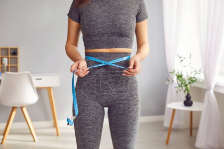 Photo for Cropped photo of faceless unrecognizable slim sporty woman wearing gray sportswear measure the waist with measuring tape standing in living room at home. Slimming and weight control concept. - Royalty Free Image