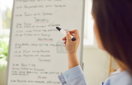 Close up of a young business woman standing in front of business planner holding marker in hand. Female worker looking to white board with weekly appointment. Planning, schedule and agenda concept.