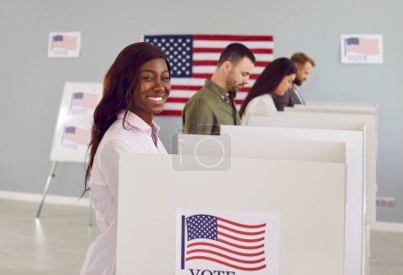 Foto de Retrato de vista lateral de una feliz mujer votante estadounidense sonriente mirando alegre a la cámara de votación y poniendo su papeleta en la papelera el día de las elecciones de pie en el centro de votación con banderas de EE.UU.. - Imagen libre de derechos