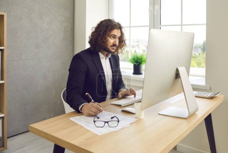 Photo for Portrait of a confident young business man accountant in suit and long hair working on modern pc computer in office typing at the desk making notes and calculations on his workplace. - Royalty Free Image