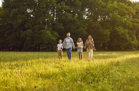 Téléchargez les photos : Bonne famille profitant de bonnes journées d'été dans la nature. Parents et enfants se promènent dans un magnifique parc ensoleillé. Maman, papa et les enfants se tenant la main et marchant ensemble sur l'herbe verte, avec des arbres en arrière-plan - en image libre de droit