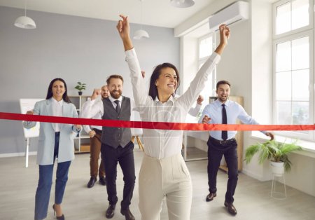 Photo for Happy woman employee running to the red ribbon with raised arms celebrating crossing finishing line in office with people colleagues in background. Business competition and teamwork concept. - Royalty Free Image