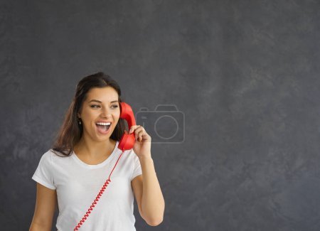 Foto de Alegre millennial gen z chica en negro estudio de fondo hablar por teléfono fijo. Sonriente joven risa tiene llamada telefónica cable agradable con el cliente. Comunicación, espacio de copia. - Imagen libre de derechos